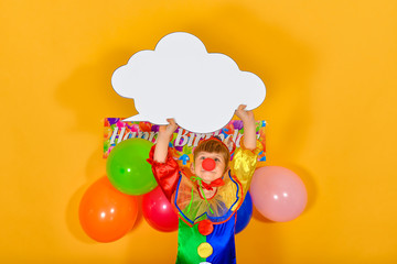 A baby clown on a birthday background, holding a white sign in the form of a cloud for inscription for inscription. Place for advertisement or announcement at a children's holiday.