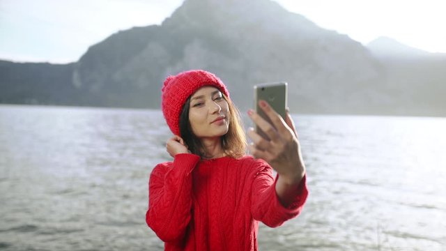 Young hipster asian woman using smartphone to take selfie in park near lake on background of Austrian Alps. Lifestyle, blogger, tourism concept.