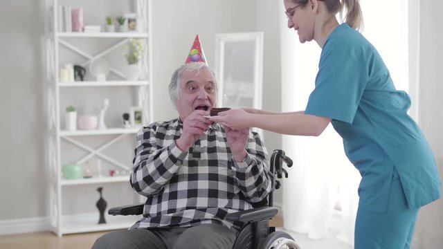 Portrait of cheerful old Caucasian man in wheelchair receiving birthday cake from invalid tender. Happy mature disabled retiree having birthday in nursing home. Happiness, lifestyle, disability, care.
