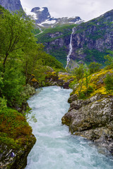 Naklejka na ściany i meble Waterfall and river which is located near path to the Briksdalsbreen (Briksdal) glacier. The melting of this glacier forms waterfall and river with clear water. Jostedalsbreen National Park. Norway.