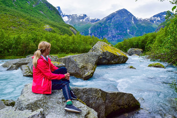 Naklejka premium The young woman near river which is located near path to the Briksdalsbreen (Briksdal) glacier. The melting of this glacier forms waterfall and river with clear water. Jostedalsbreen National Park. No