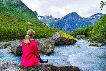 The young woman near river which is located near path to the Briksdalsbreen (Briksdal) glacier. The melting of this glacier forms waterfall and river with clear water. Jostedalsbreen National Park. No