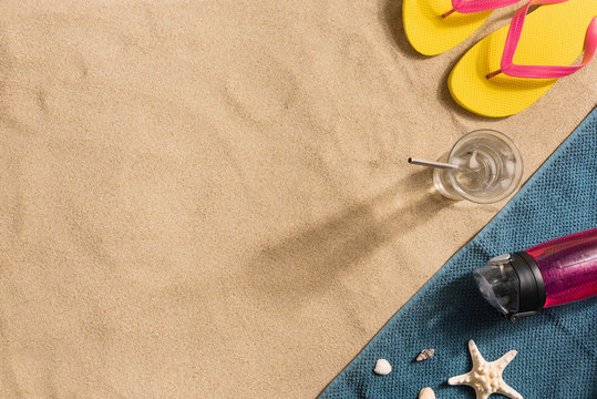Summer vacation composition. Flip flops, water bottle and glass of water on sand background. Harsh light with shadows. Summer background. Border composition made of towel