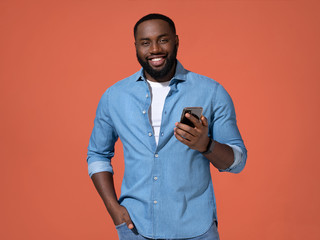 Handsome man using smartphone. Photo of african man in casual outfit on coral background.