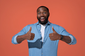 Happy man shows thumbs up with both hands. Photo of african man in casual outfit on coral background.