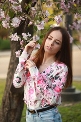 Obraz premium A woman dressed in a delicate shirt with a raspberry flower print and blue trousers with a blossoming apple tree in spring.