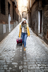 Fototapeta na wymiar young girl sightseeing through the streets of a city with yellow coat, wool cap and suitcase