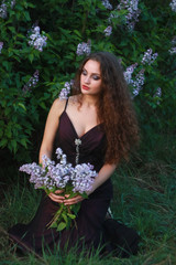 A woman dressed in a beautiful purple dress on a background of lilac bushes with a bouquet of lilac in the hands of spring.
