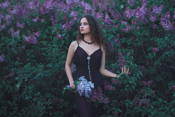 A woman dressed in a beautiful purple dress on a background of lilac bushes with a bouquet of lilac...