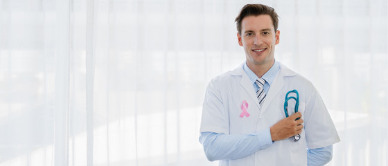 healthcare background of caucasian male doctor holding stethoscope on hand with pink ribbon on lab coat in concept of fighting against cancer