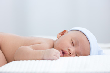 A 1-month-old infant is wearing a blue hat is sleeping on the white futon. The rest of the newborn baby.