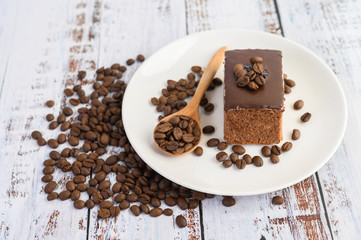 Chocolate cake on a white plate and coffee beans on a wooden spoon on a wooden table.
