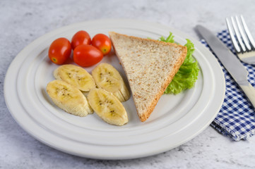 bread, banana, and tomato on white plate with fork and a knife.