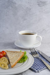 bread, banana, and tomato on white plate with coffee cup, fork, and a knife.