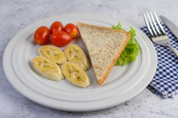bread, banana, and tomato on white plate with fork and a knife.