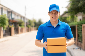 Asian delivery young man in blue uniform smile and holding pile of cardboard boxes in front house village with copy space. Advertising, Business, Transportation Concept..