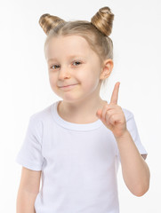 The little blonde smiles slyly and shows her index finger in a white t-shirt on a white background. The concept of an idea or attracting attention.