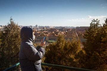 Picture of Chinese girl wearing mask to protect corona or covid-19 virus and playing phone with city view of forbidden city, Beijing, China and dark blue sky as a background.