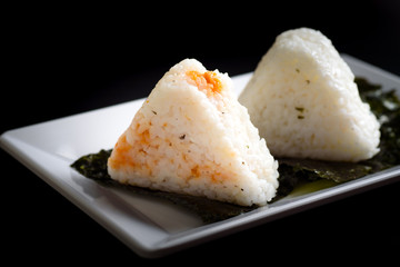 Onigiri, a Japanese food that looks like a triangle or ball oval Wrapped in seaweed (Nori), is a healthy food placed on a white plate with a black background