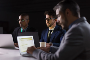 Confident men working over project in office late at night. Thoughtful business team sitting at table with laptops. Teamwork, working late concept