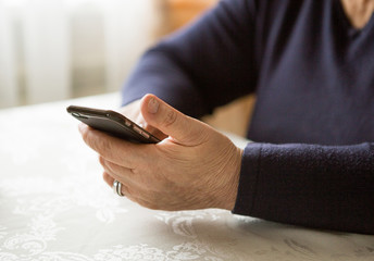 Retired woman is using mobile phone,  at cozy home kitchen. Grandmother has coffee break in cooking. Senior person is looking for recipe, communication online in the internet. Authentic moment.