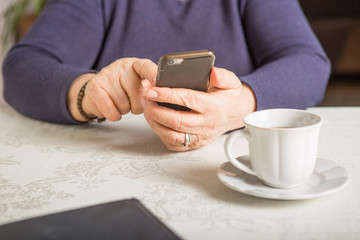 Retired woman is using mobile phone,  at cozy home kitchen. Grandmother has coffee break in cooking. Senior person is looking for recipe, communication online in the internet. Authentic moment.