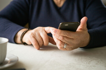 Retired woman is using mobile phone,  at cozy home kitchen. Grandmother has coffee break in cooking. Senior person is looking for recipe, communication online in the internet. Authentic moment.