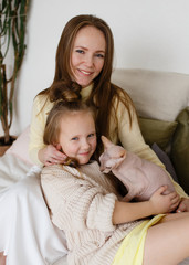 The concept of family photography. Mother and daughter in cozy home clothes playing with a pet, with a cat sitting on the sofa in a cozy living room decorated with plants, happiness joy love care