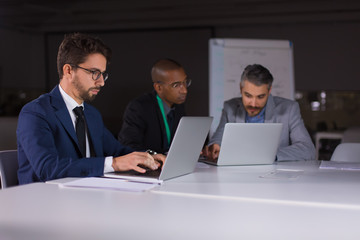 Group of confident businessmen working with laptops in office. Thoughtful young managers working together in office at night. Teamwork, working late concept
