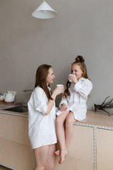 Obraz na płótnie Canvas Happy mother and daughter in home white shirts relax in a hammock, have Breakfast and drink tea in the kitchen, have fun, communicate, hug in a cozy bright house, comfort, love, care, family, nature