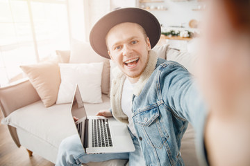 Happy hipster man smile freelancer traveler taking selfie photo in hat, working on laptop home. Concept travel ticket booking