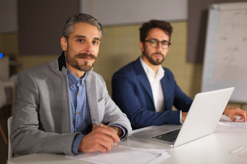 Two handsome businessmen looking at camera. Cheerful bearded manager sitting at table and holding pen. Business, working late concept