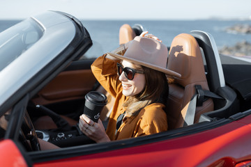 Young stylish woman in hat driving convertible car near the ocean, enjoying summer vacations,...