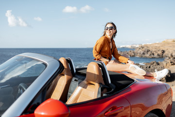 Portrait of an attractive woman in swimsuit and shirt sitting on the car hood, while traveling by car near the ocean. Concept of a summer time and wellbeaing