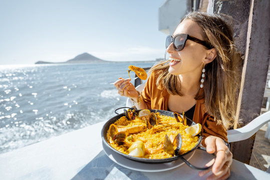 Woman eating paella, traditional spanish dish, while sitting at the restaurant terrace near the ocean. Concept of sea food and good summer vacations