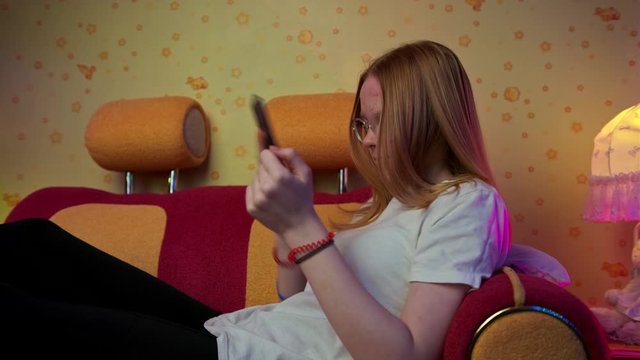 lovely schoolgirl shakes lagging smartphone sitting on bright red and orange sofa against pretty wallpaper closeup