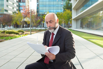 Concentrated businessman reading reports outdoors. Focused bearded mature man sitting on street...