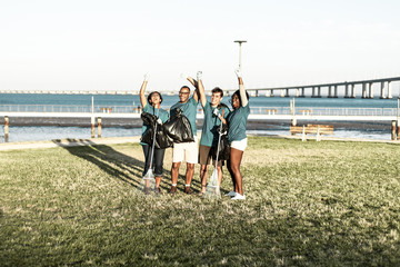 Group of happy volunteers celebrating success outdoors. Mix raced men and women wearing uniforms, standing on grass, holding rakes and plastic bags and making winner gestures. Success concept