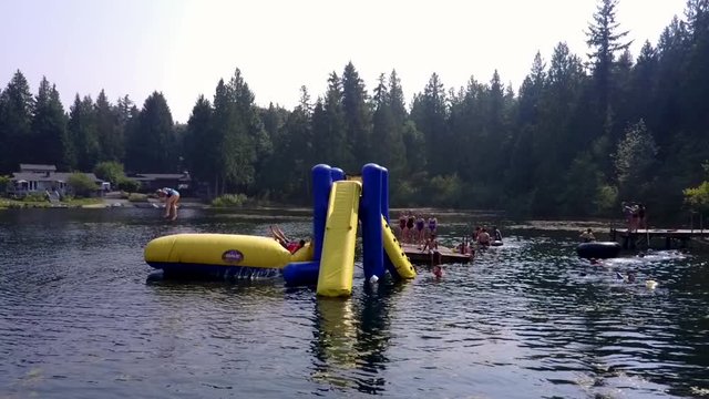 A kid jumps onto an inflatable air cushion and sends their friend flying into the air and then into a lake in a slow motion, drone shot.