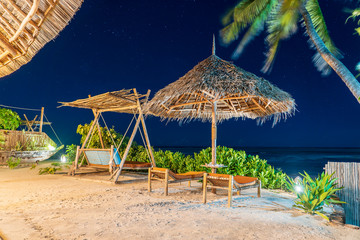 Wooden swing with a mattress under a canopy and straw umbrella on the tropical beach near sea at night, island Zanzibar, Tanzania, Africa