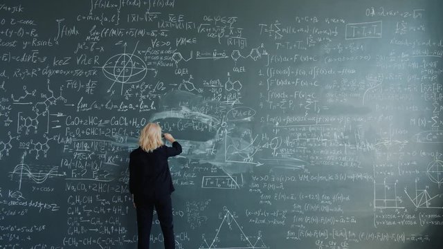 Smart woman researcher is walking to blackboard in classroom writing formulas with chalk focused on science theory. People, education and work concept.