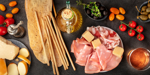 Italian food panorama, antipasti flat lay. Charcuterie, cheese, capers, wine, tomatoes, grissini, shot from the top on a black background