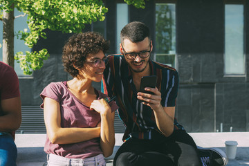Cheerful geek couple discussing online mobile app. Man and woman in glasses sitting on parapet...