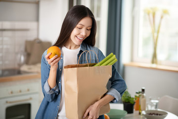 Pretty woman with orange in hand looking into bag with vegetables.