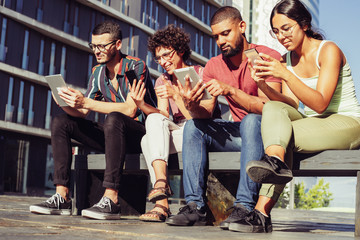 Group portrait of young people focused on their digital gadgets. Men and women sitting on bench outside, using mobile app or consulting internet. Communication concept