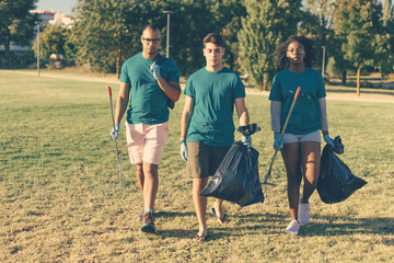Serious volunteers carrying garbage from city park. Young woman and men walking through city lawn, holding rakes and plastic bags. Trash removal concept