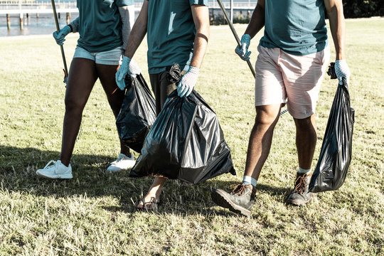 Volunteers cleaning park area from rubbish. Young woman and men walking on grass, carrying rakes and full plastic bags. Waste collection concept