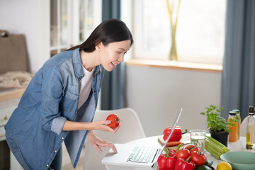 Stylish young girl standing near laptop with tomato in hand.