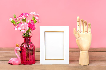 Small colored pink carnations in vase, white photo frame, figure of flamingo, wooden hand on wooden table and pink wall Hello spring, seasonal concept Mock up Place for text