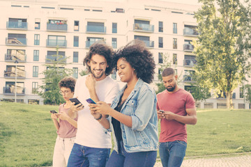 Happy mix raced couple using smartphones and showing content to each other. Man and woman with mobile phones walking behind them. People with gadgets concept
